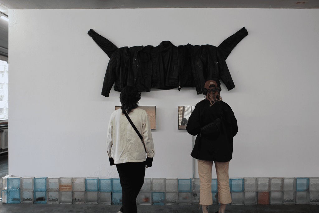 Zwei Personen stehen vor einem Kunstwerk, dieses besteht aus zwei Bildschirmen, darüber hängt eine Installation aus schwarzen Lederjacken.